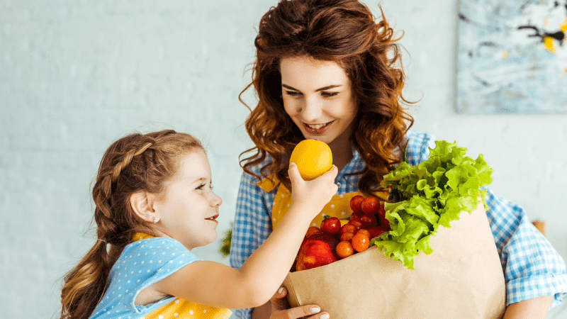 A young girl takes an orange out of a groceries bag as her mom smiles.