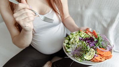 Pregnant woman eats a variety of vegetables from a large plate.