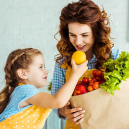 A young girl takes an orange out of a groceries bag as her mom smiles.